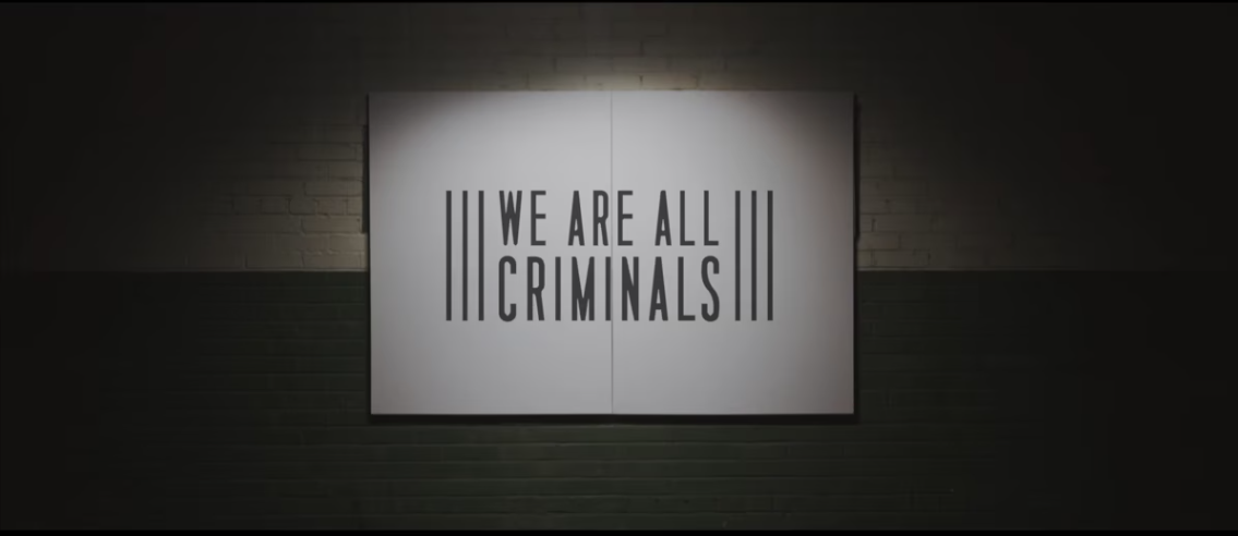 We Are All Criminals Black logo on white sign leaning against brick wall