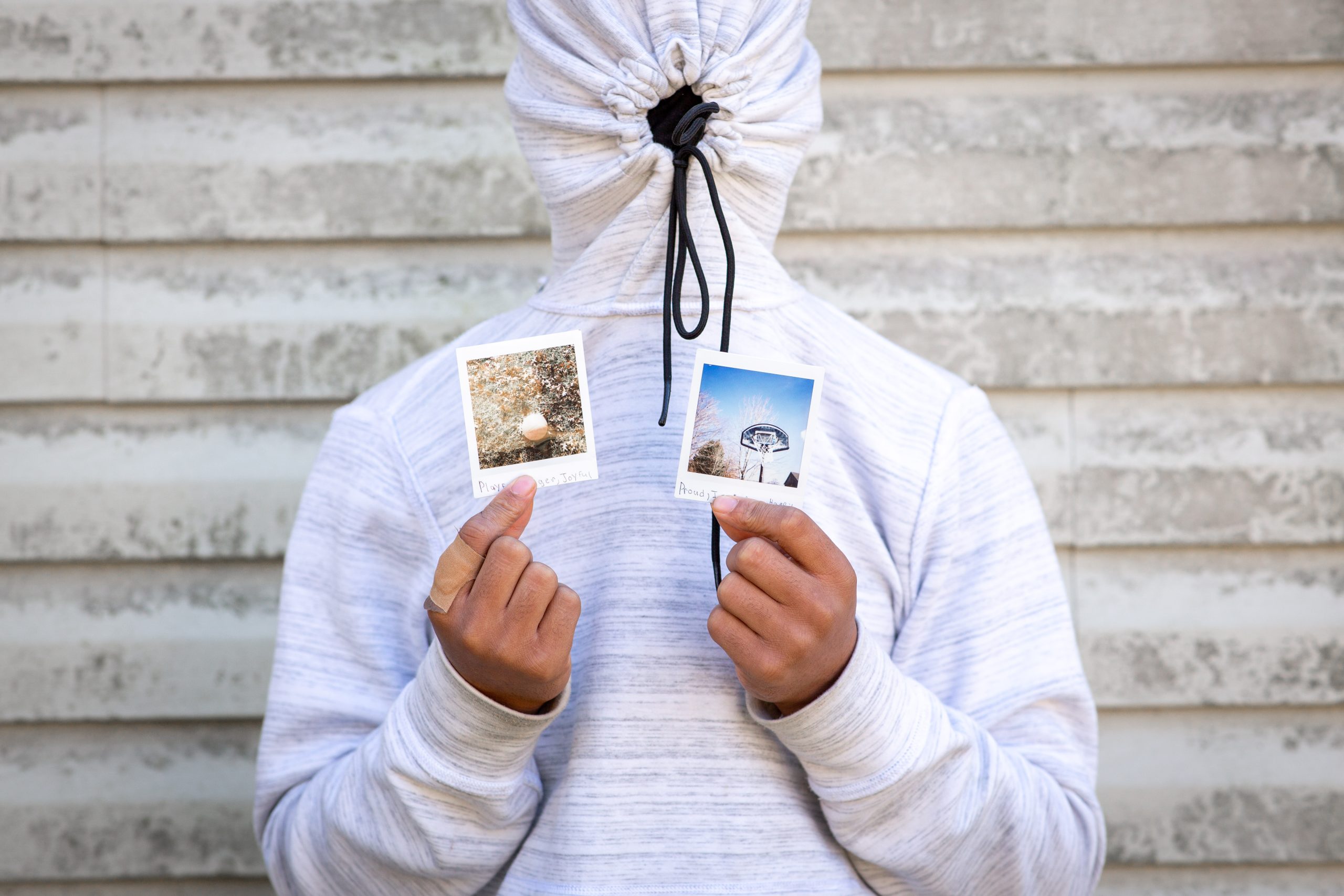 young man in white hoodie, with strings pulled tight over his face. holding two polaroids