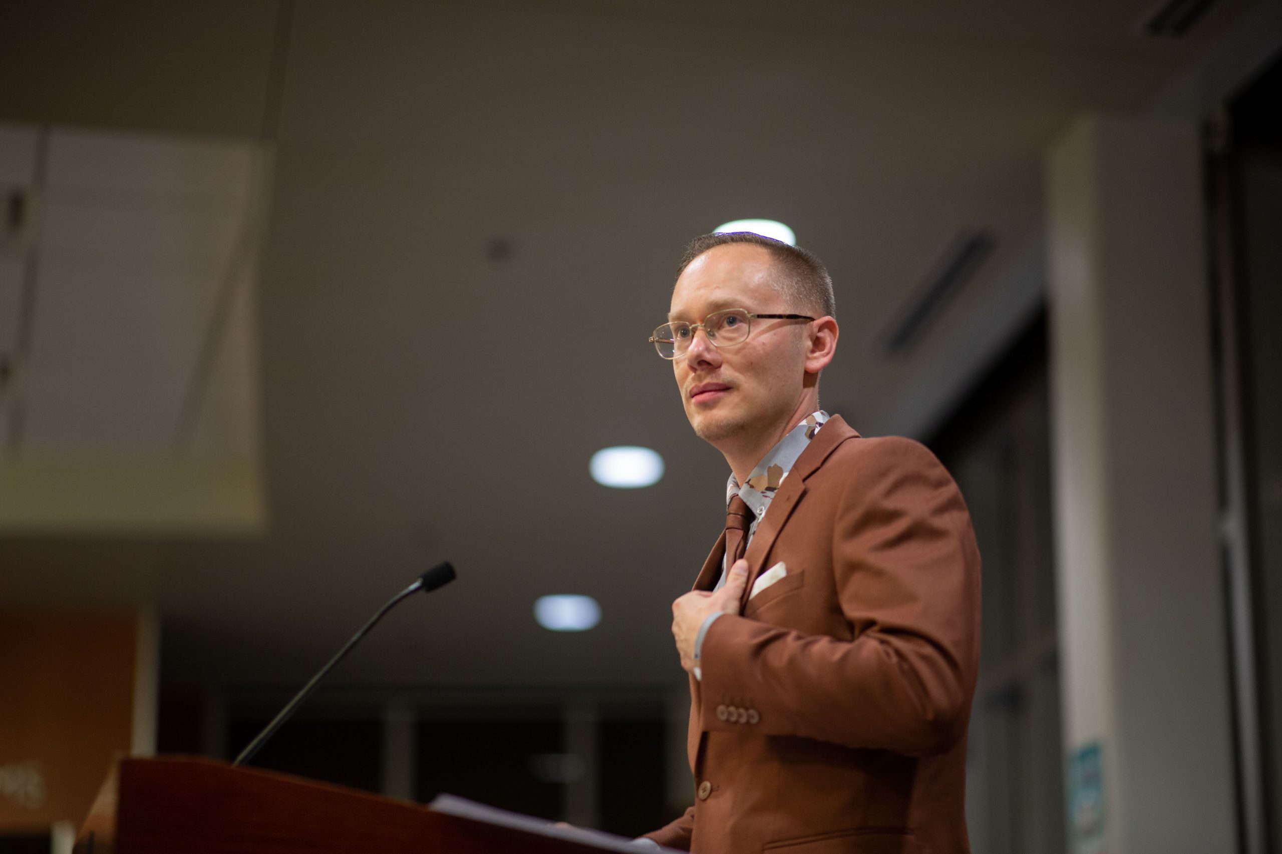 Poet and memoirist Zeke Caligiuri at the mic, in a brown suit with his left hand resting on his chest
