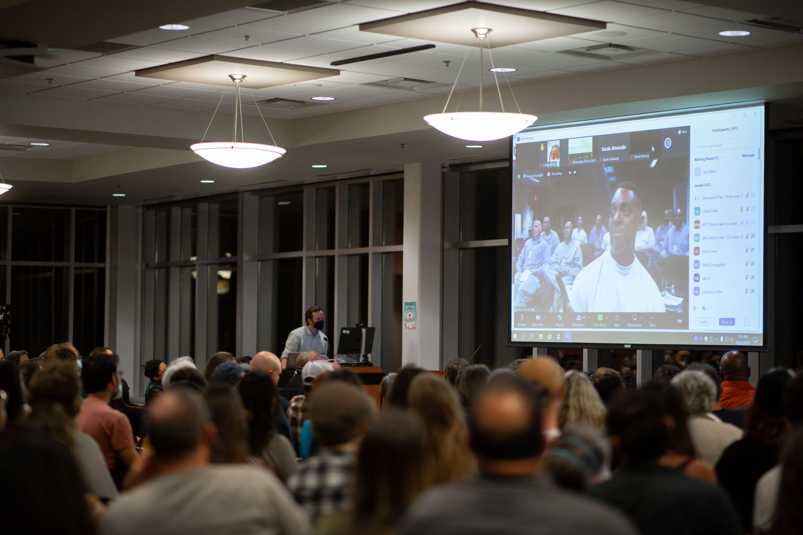 Poet Bino appears on a large screen, appearing in to the conference space via Zoom; the room is full of people, watching and listening to him