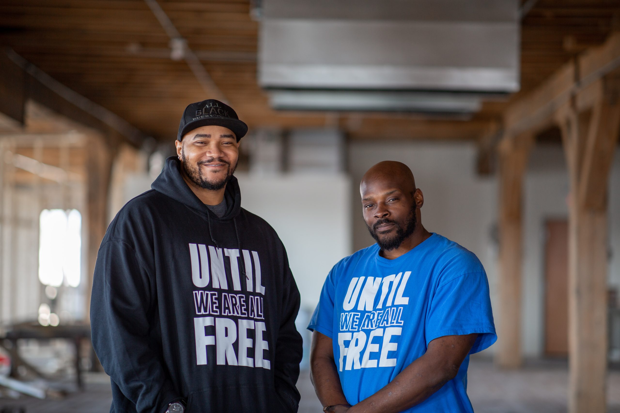 Two people in Until We Are All Free shirts, standing in warehouse, looking directly at camera
