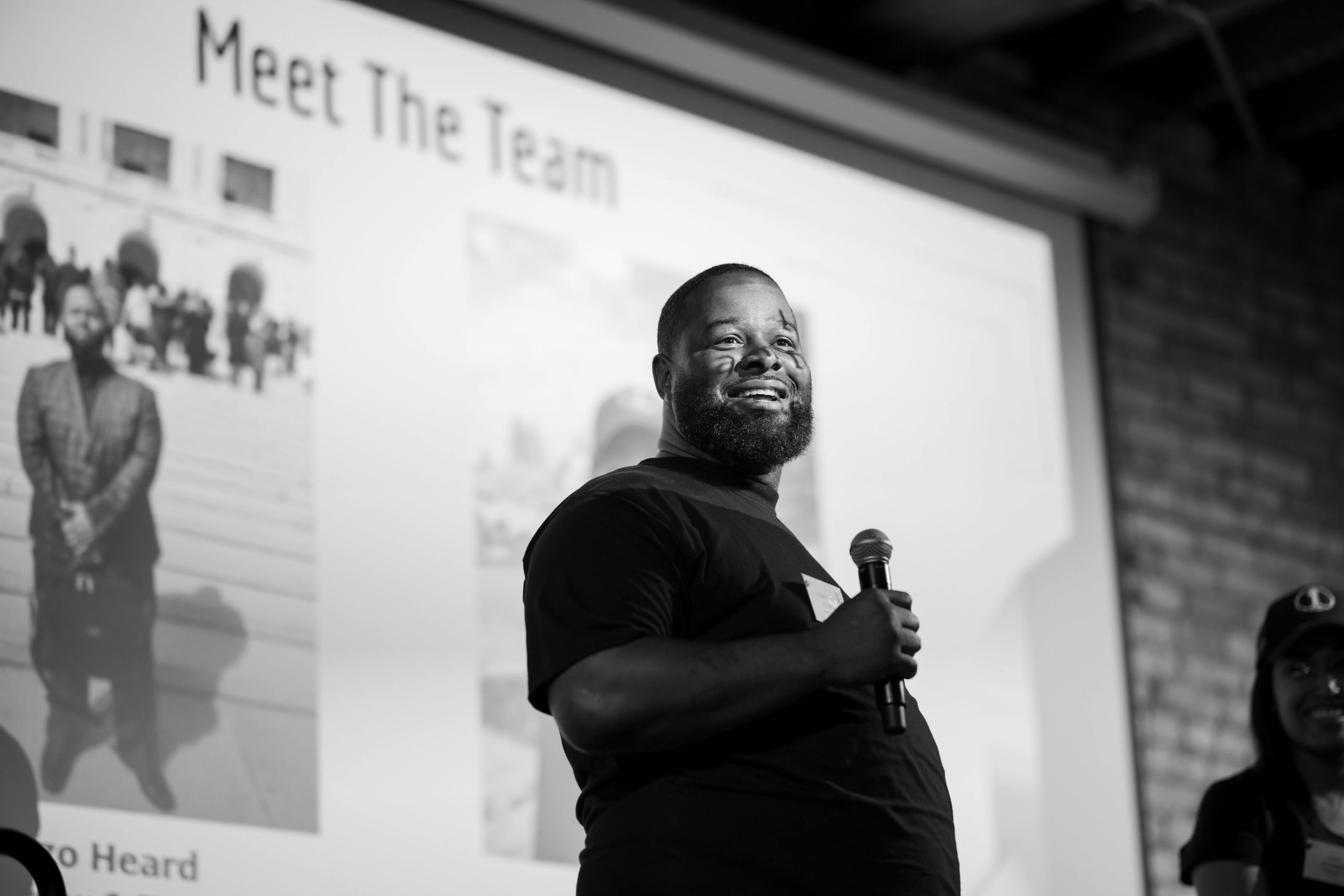 Lorenzo Heard, smiling and holding a microphone and standing in front of a powerpoint that says "Meet the Team"