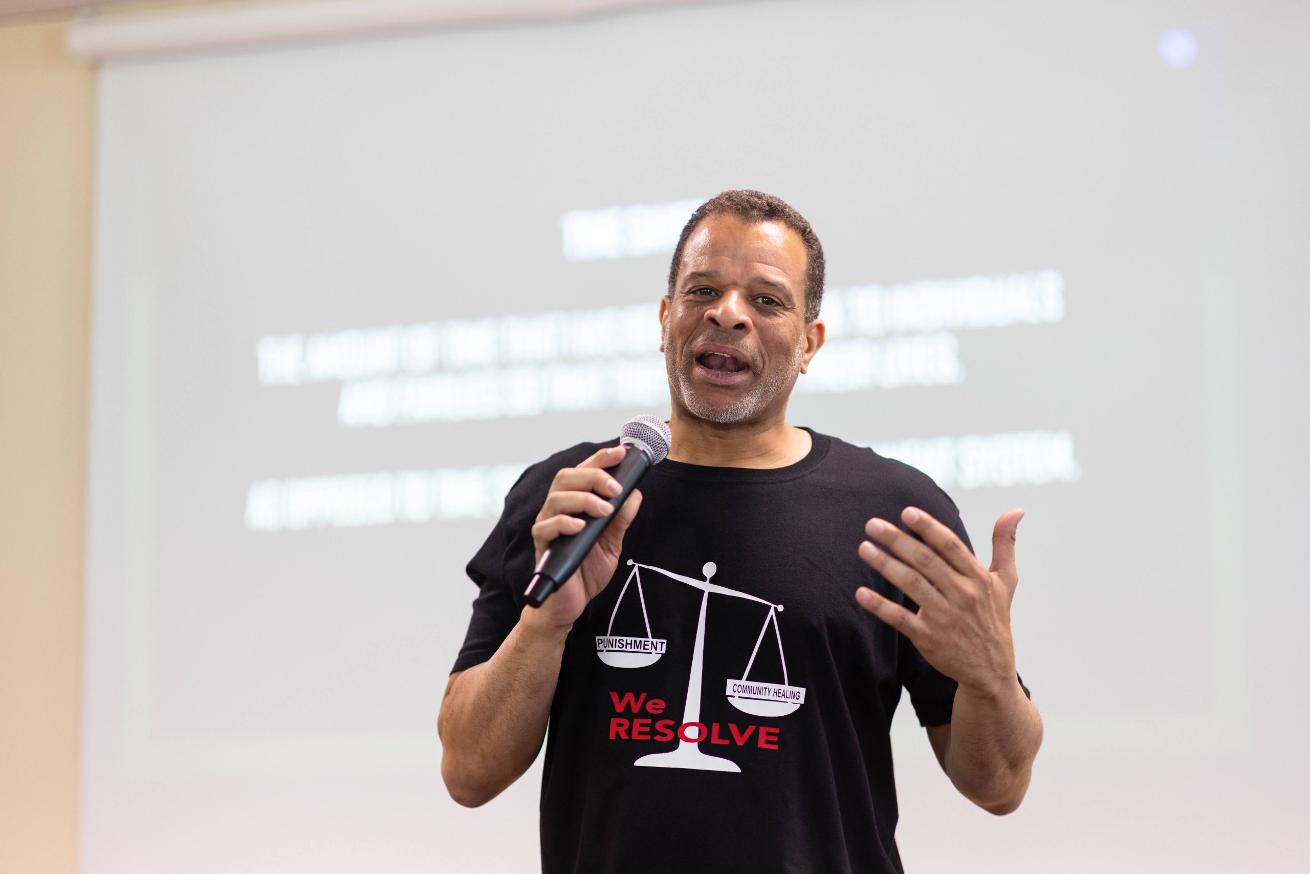Damon at the mic at a We Resolve event, powerpoint in the background