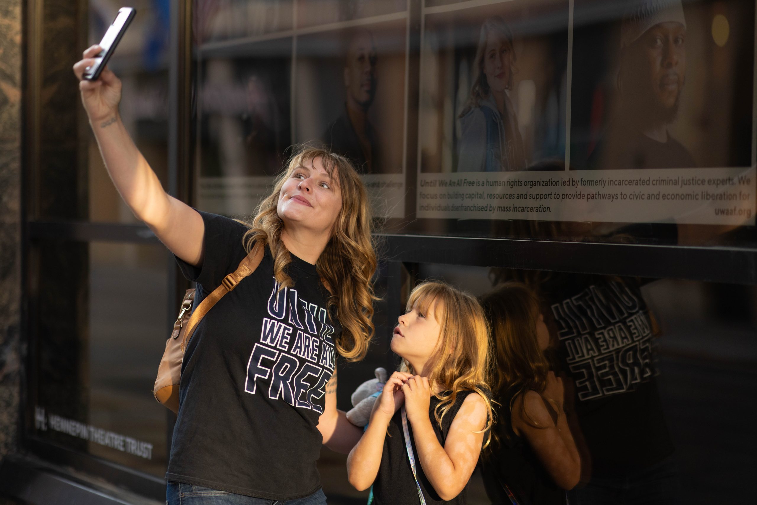Kahlee and daughter in front of Kahlee's portrait in the Hennepin Theatre Trusts' sidewalk exhibit. She is taking a selfie and looking toward the sun