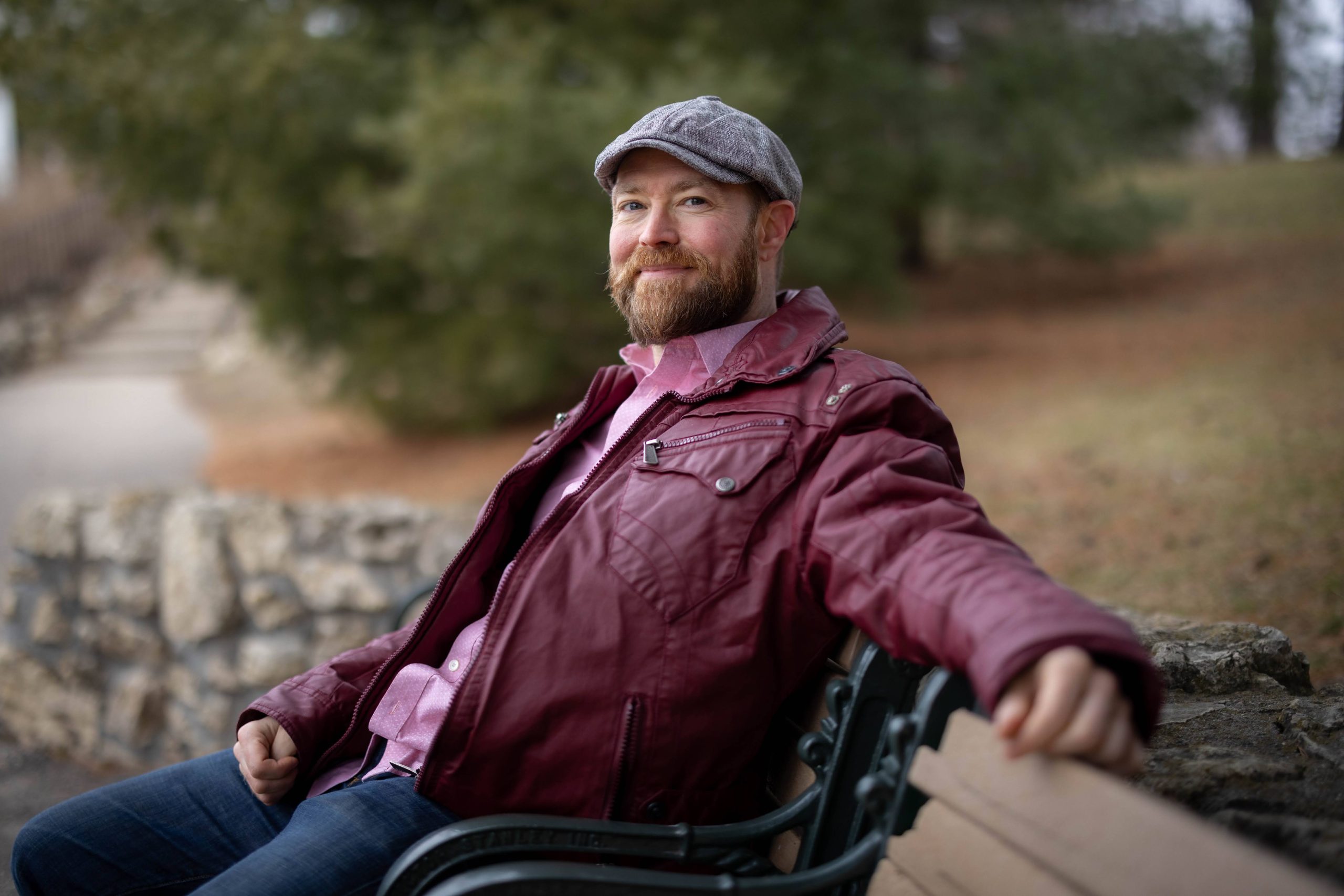Artist Luke on a park bench, wearing a grey hat and a purple jacket, smiling at the camer