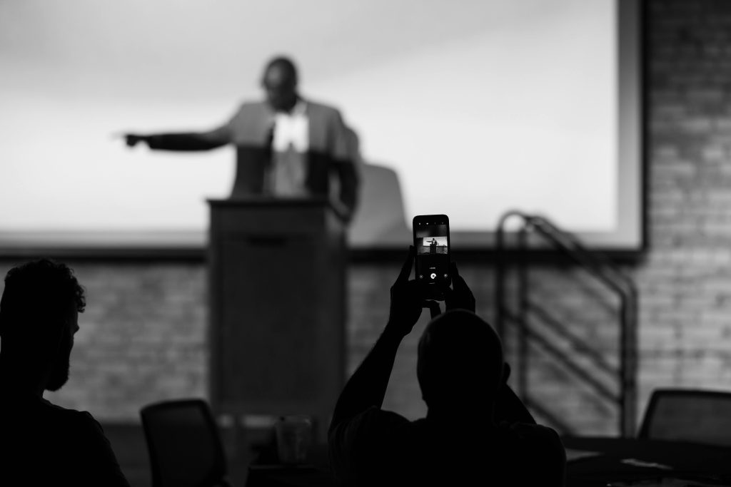 Black and white photo, focused on an audience member's phone's screen, recording Senator Bobby Joe Champion, speaking and gesticulating on stage.