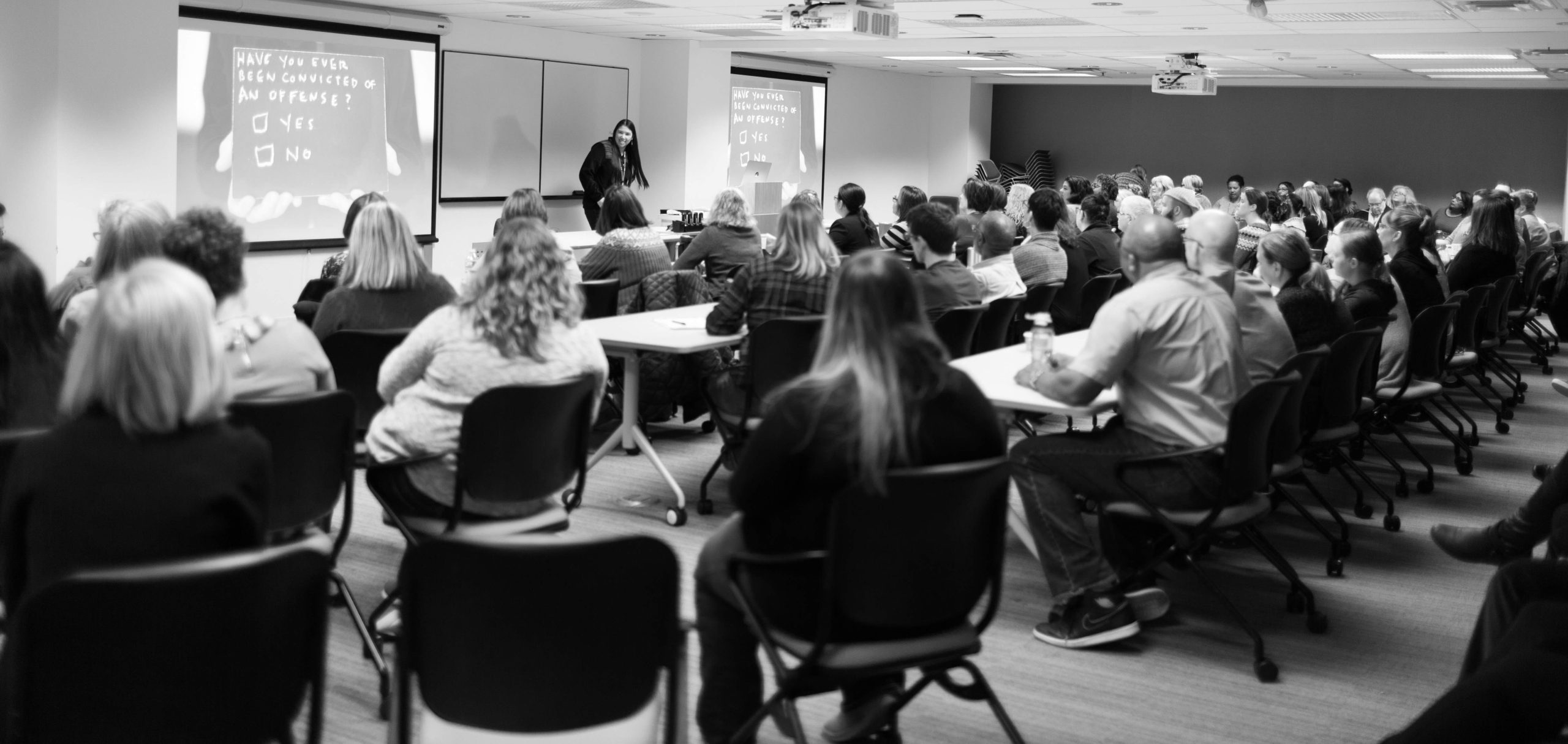 Alicia presenting to a group of county employees in Ramsey. Image is in black and white, with Alicia at the front of the room smiling, a powerpoint presentation on the screen in the background.