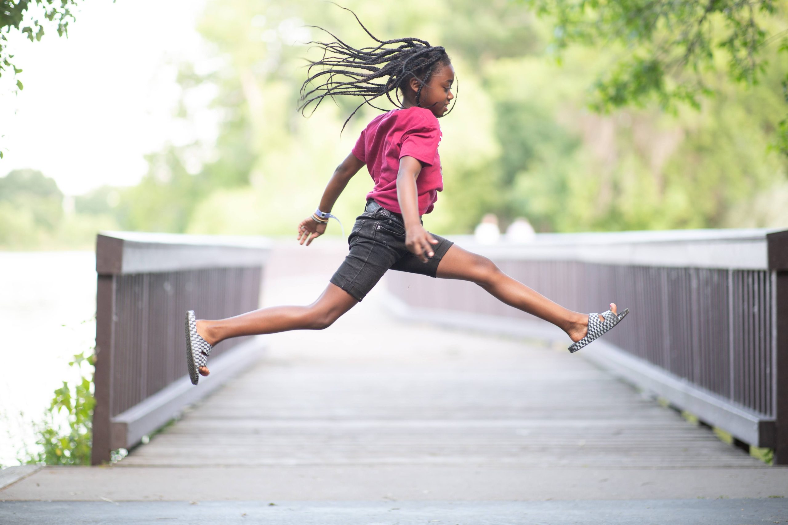 Young Black girl in braids leaping in front of wooden bridge