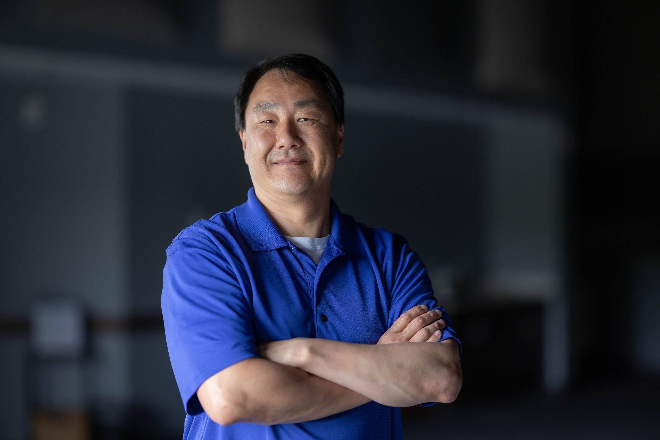Man in bright blue shirt, turned toward camera with arms crossed in front of chest, smiling